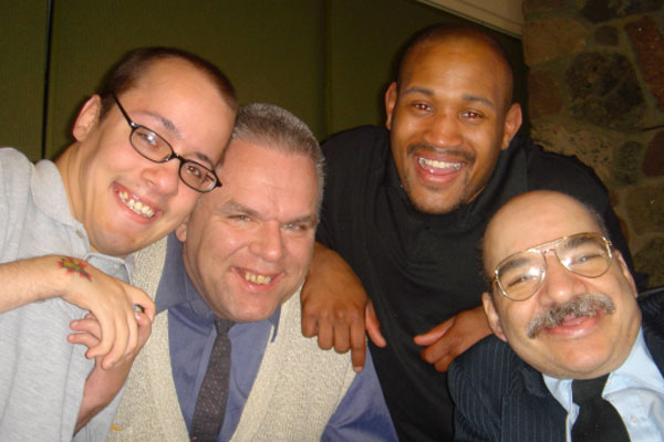 Four happy adults of diverse backgrounds in a close embrace, smiling at the camera in a warm, friendly setting